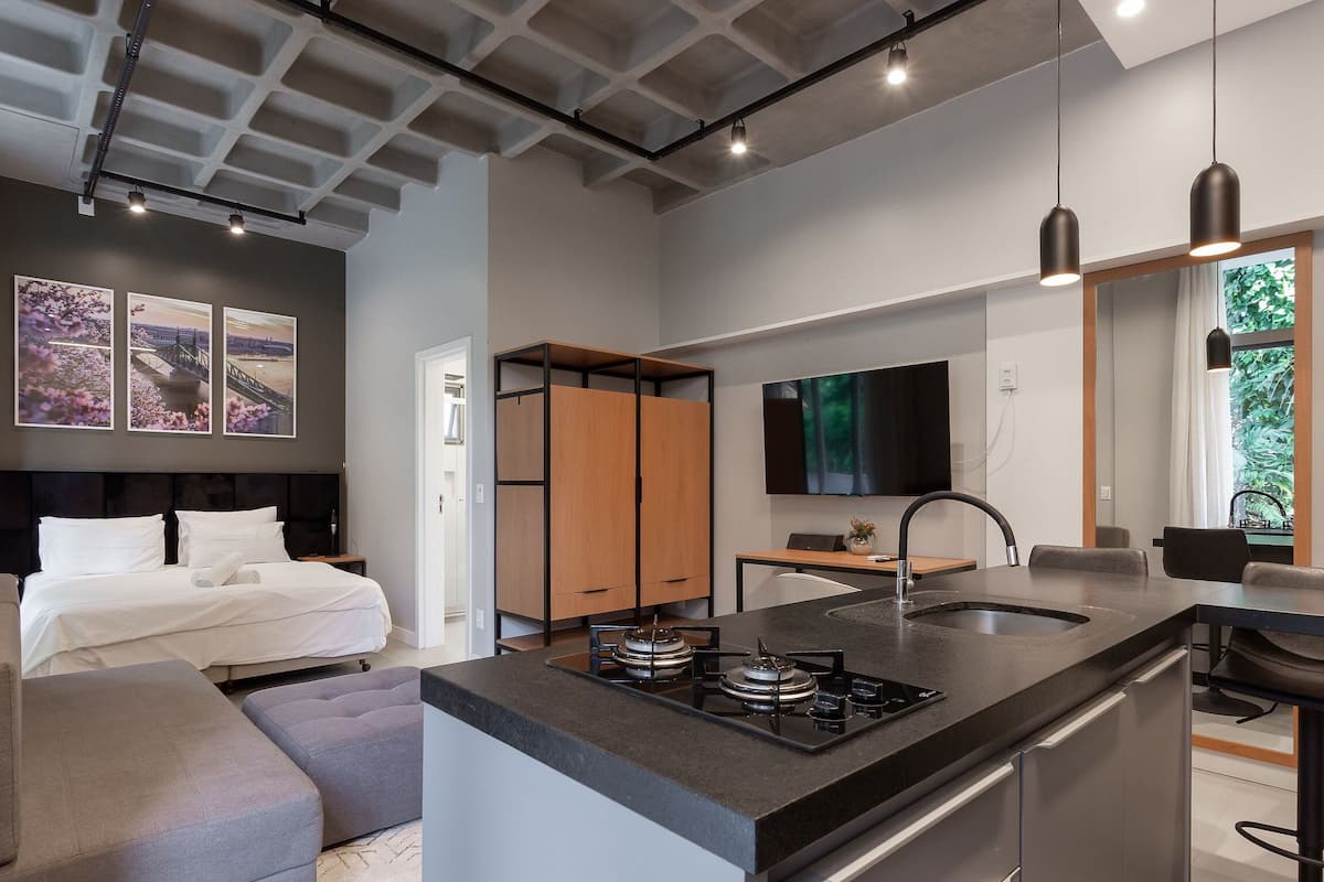 A modern hotel room with a bed, a sofa, a kitchenette with a stove, a TV, and artwork on the walls.