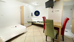 Una habitación pequeña y bien iluminada con cama, escritorio, sillas, armario y televisor montado en la pared.