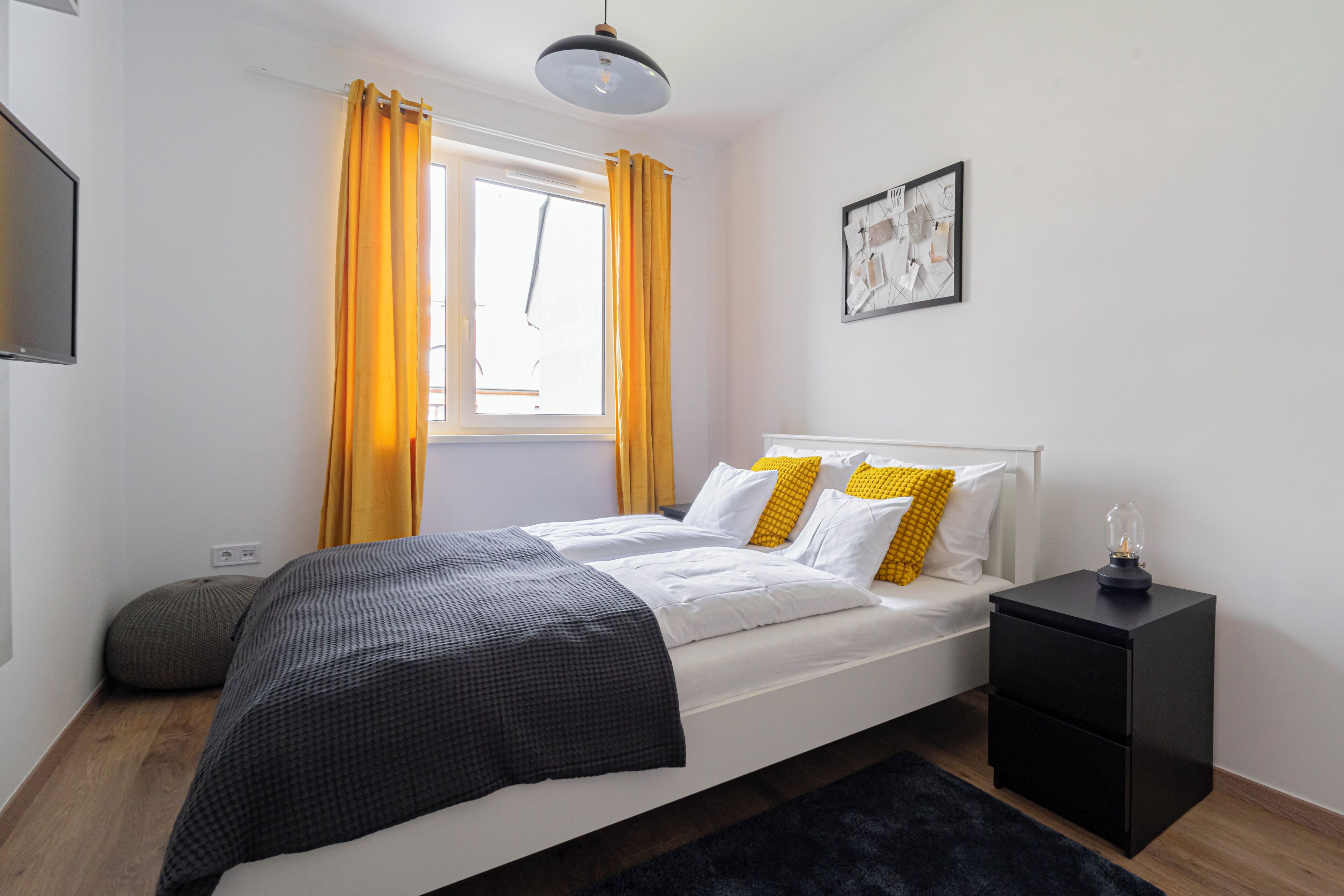 A bedroom with a bed, a nightstand, a window with curtains, a wall-mounted TV, and a framed picture.
