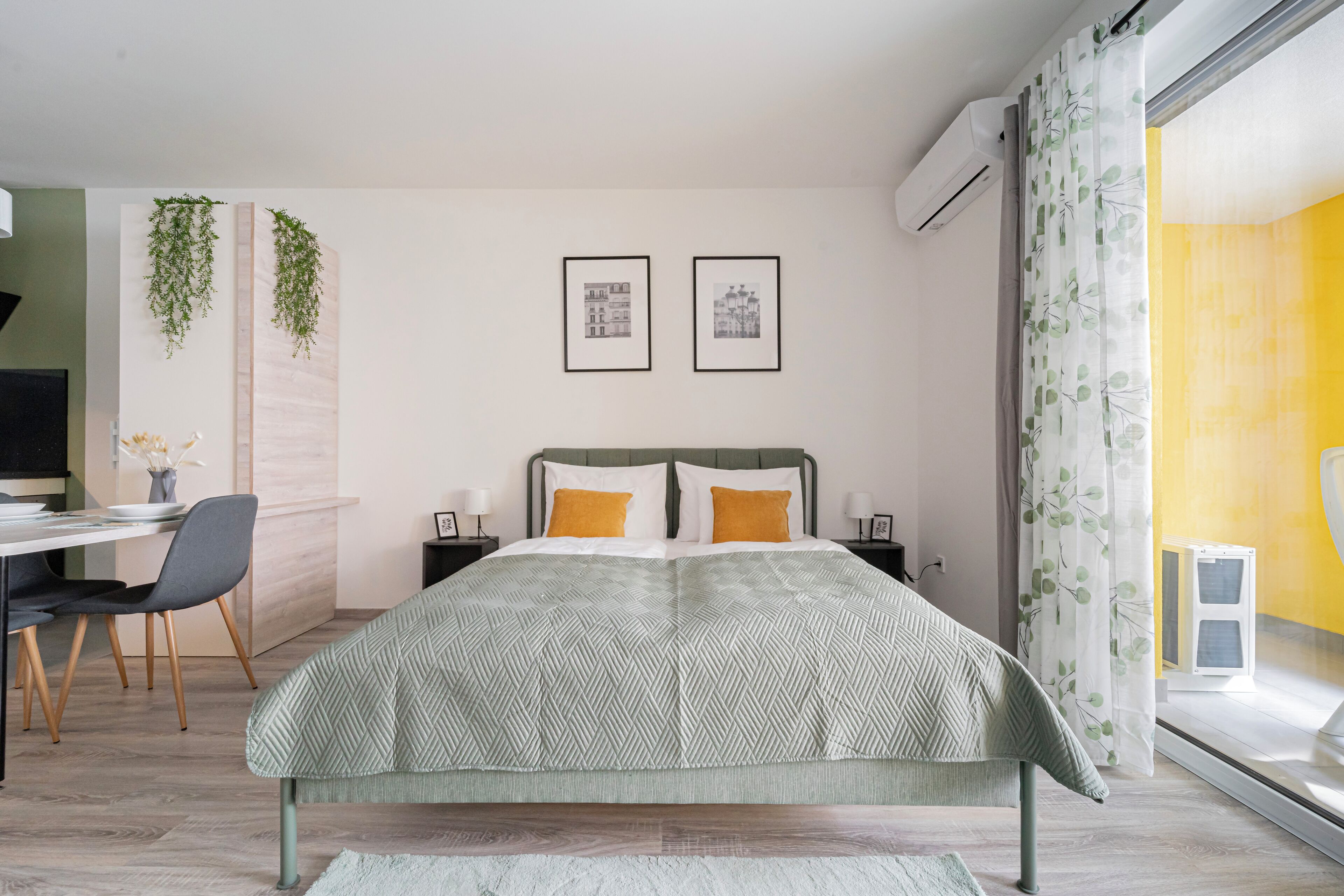 A modern bedroom with a bed, a desk, and a chair. There are two framed pictures on the wall, a plant, and an air conditioning unit.