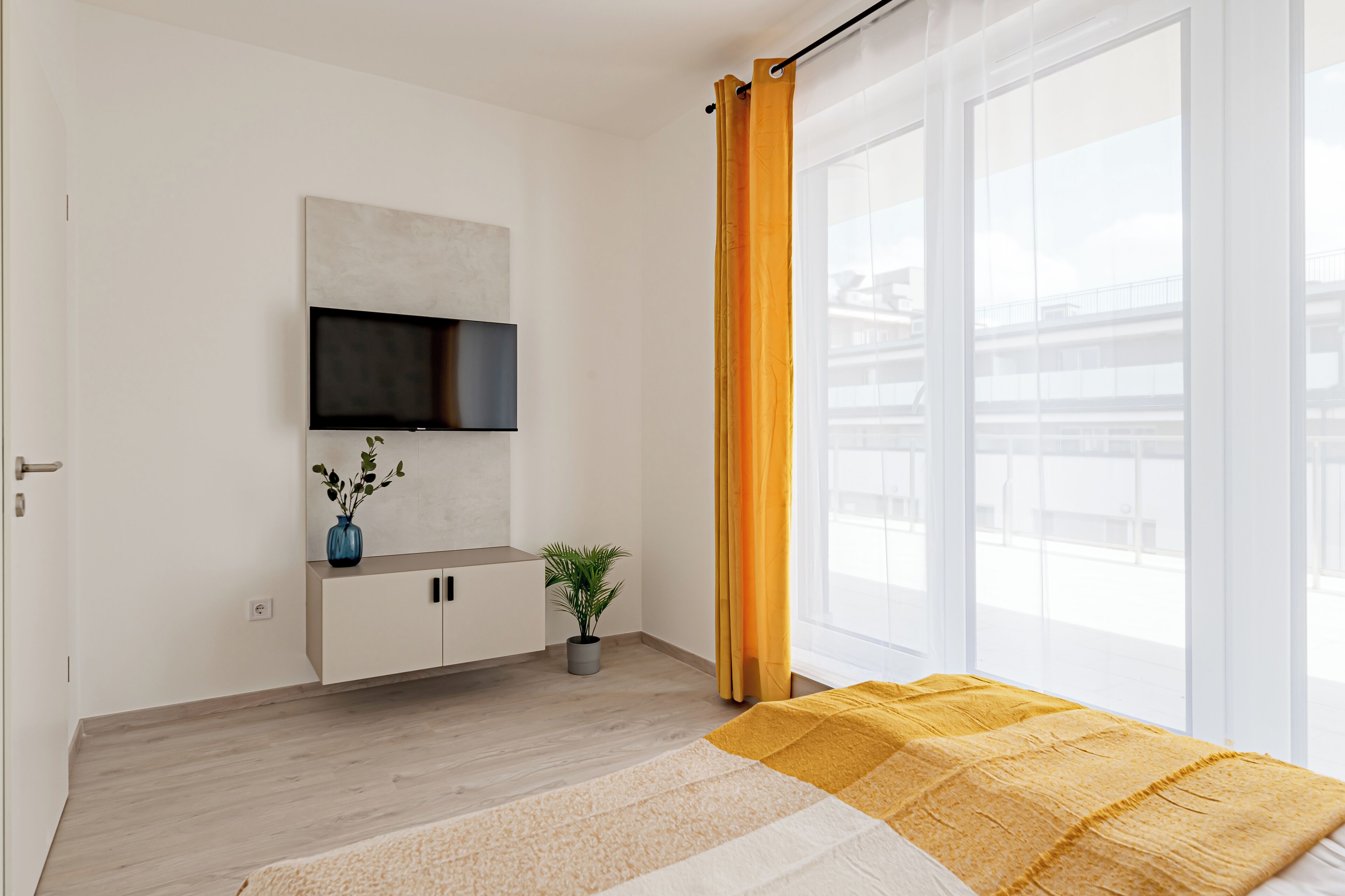 A modern hotel room with a large window, a flat-screen TV mounted on the wall, a small potted plant, and a bed with a yellow blanket.