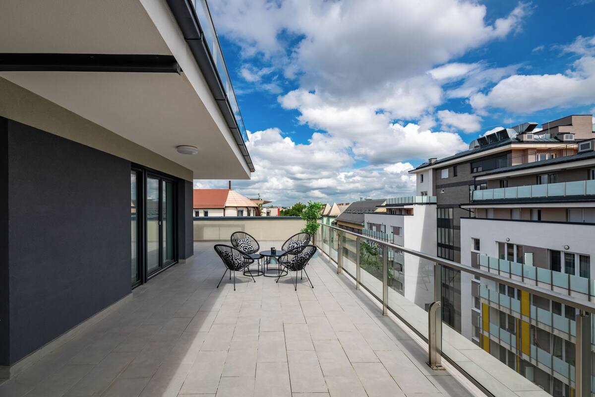 A modern balcony with a glass railing, a table with chairs, and a view of surrounding buildings.