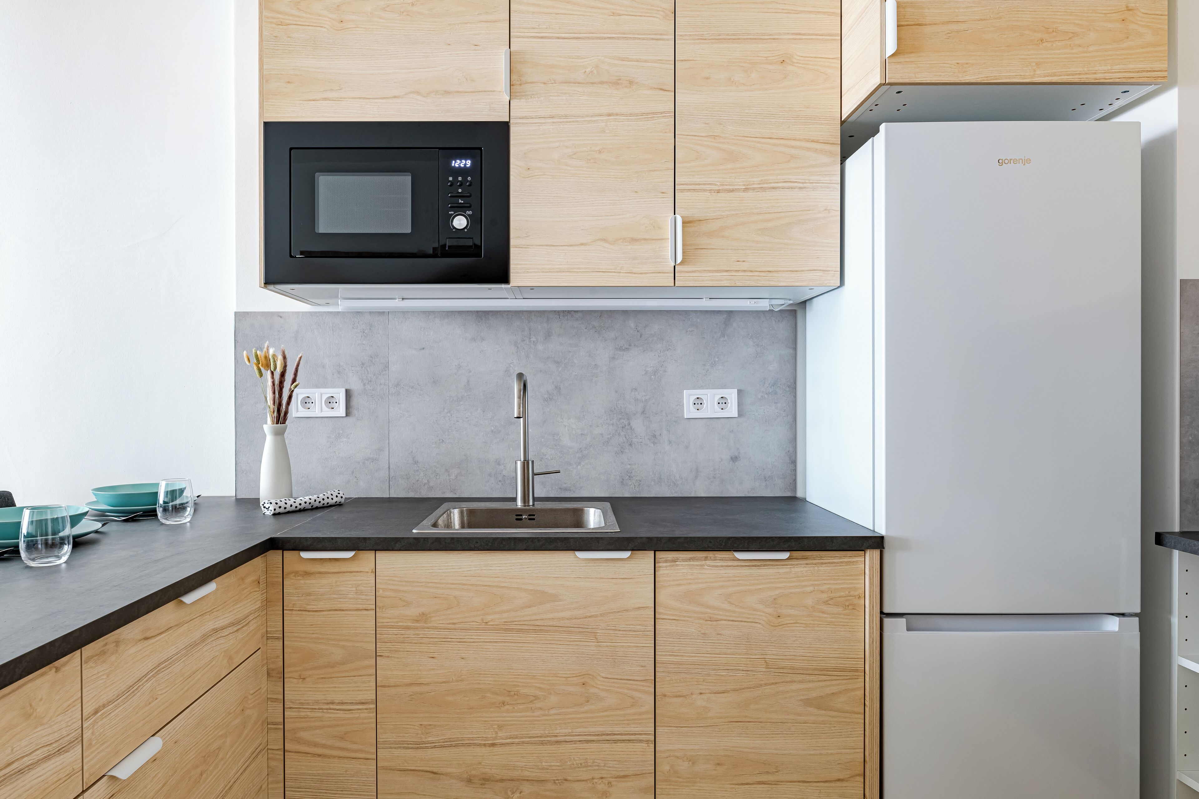 A modern kitchen with wooden cabinets, a black microwave, a white refrigerator, and a grey countertop.