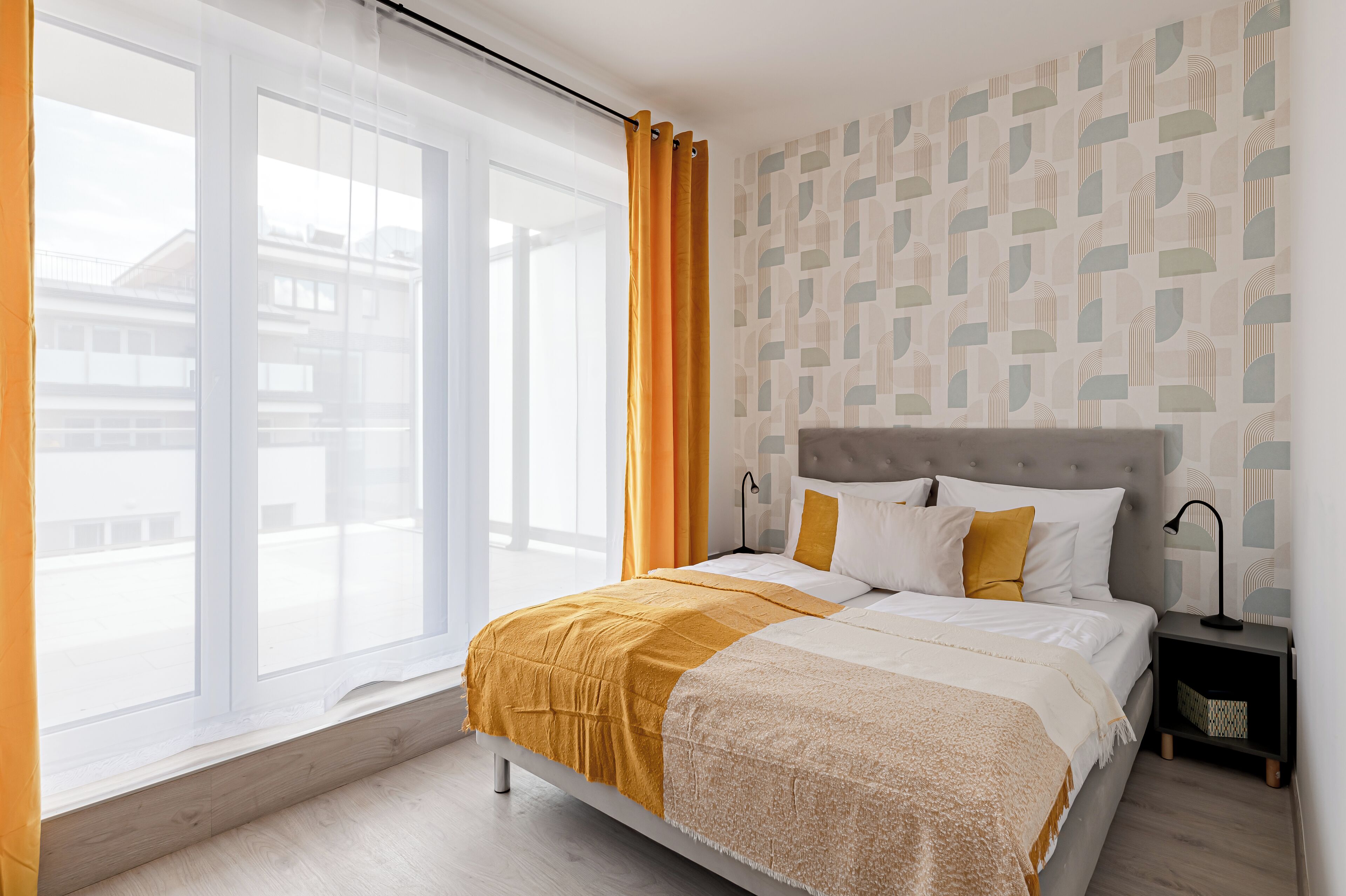 Une chambre avec une grande fenêtre, un lit recouvert d’une courtepointe blanche et jaune, une tête de lit grise et un mur à motifs.