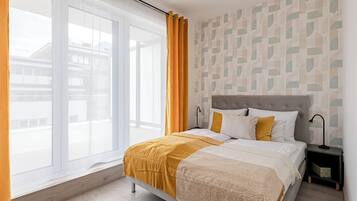 Une chambre avec une grande fenêtre, un lit recouvert d’une courtepointe blanche et jaune, une tête de lit grise et un mur à motifs.