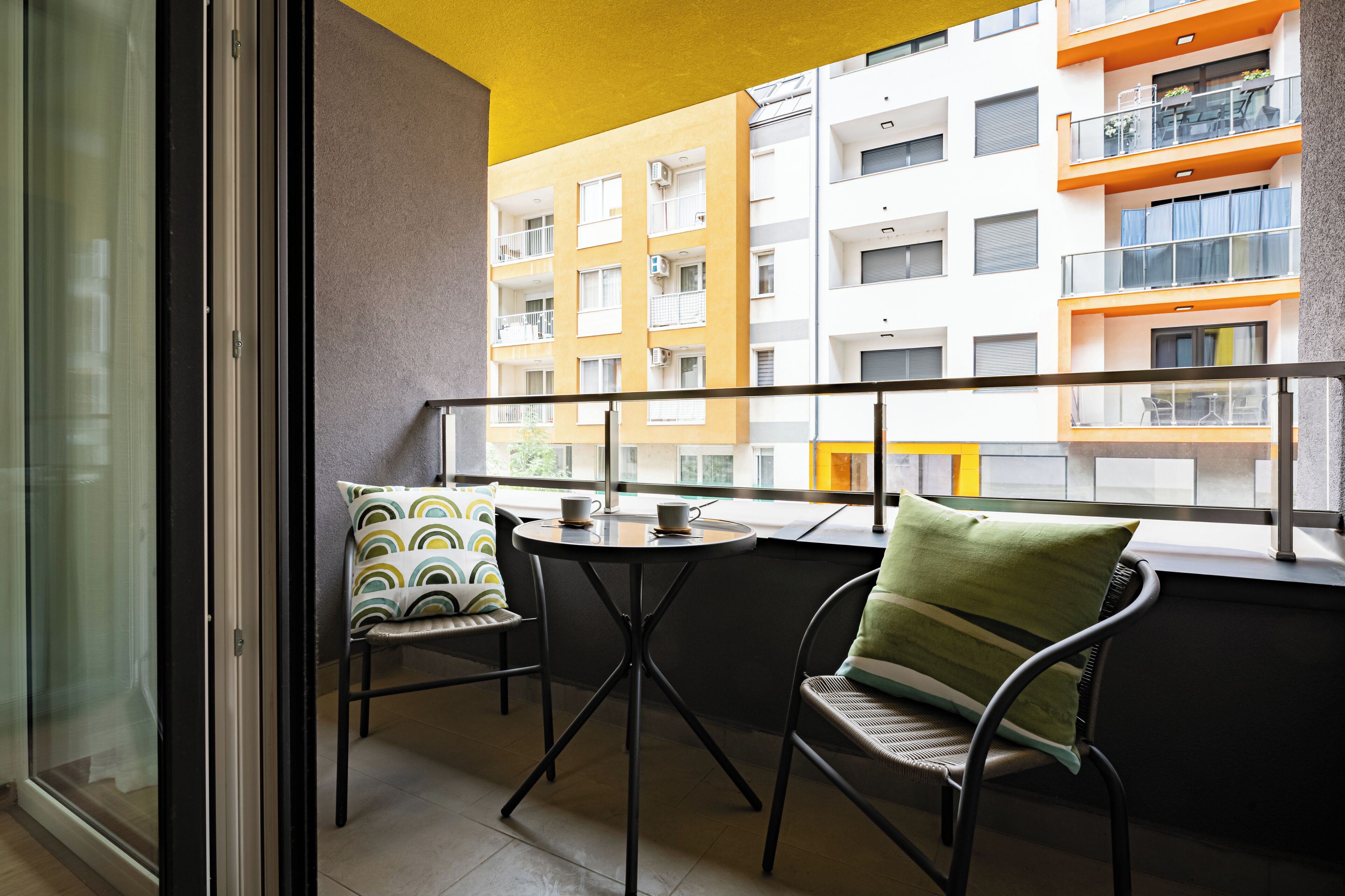 Un balcón con mesa y sillas, vista a un edificio de varios pisos también con balcones, y un techo amarillo.