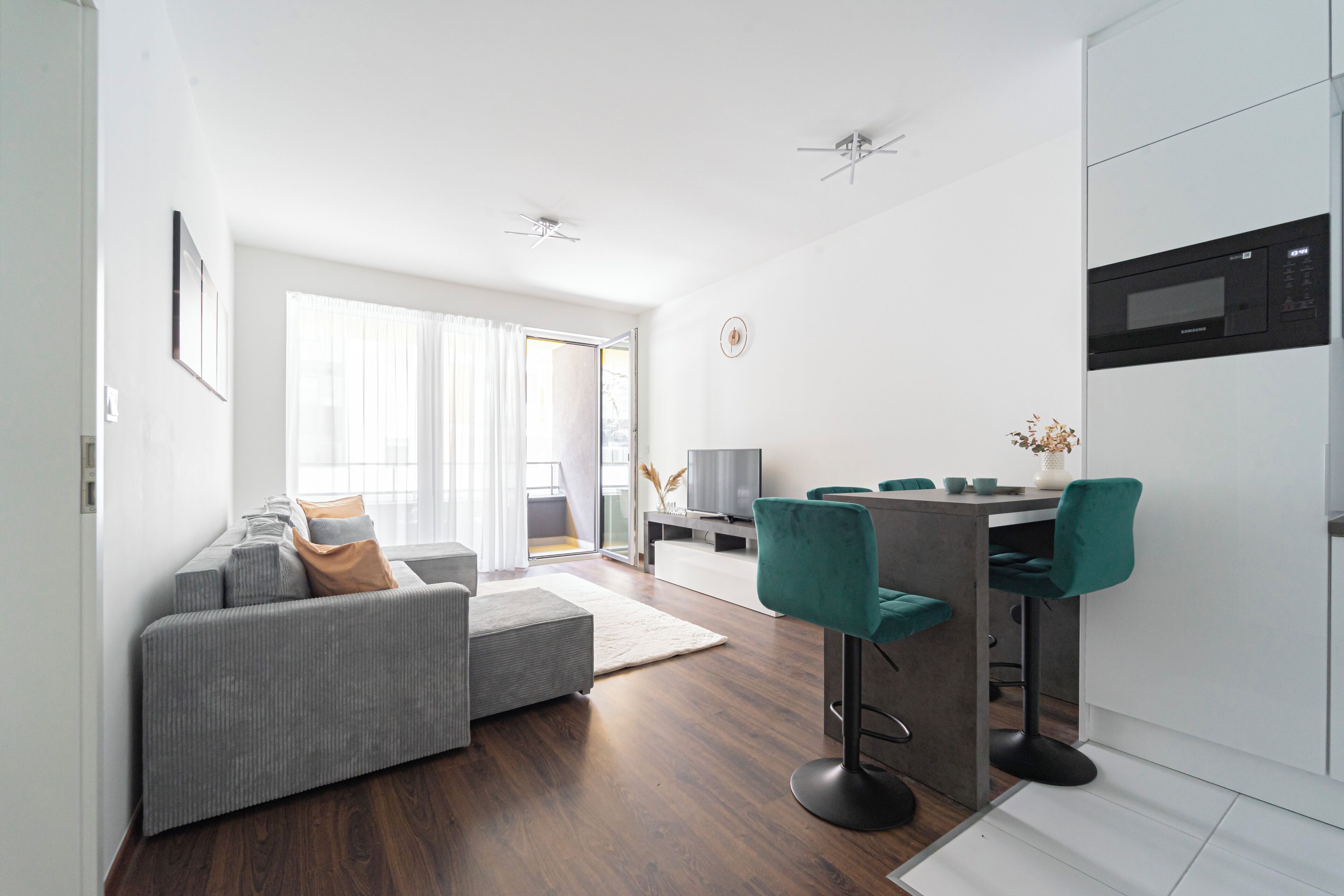 A modern living room with a grey sofa, teal bar stools, and a dining area with a white table and chairs.