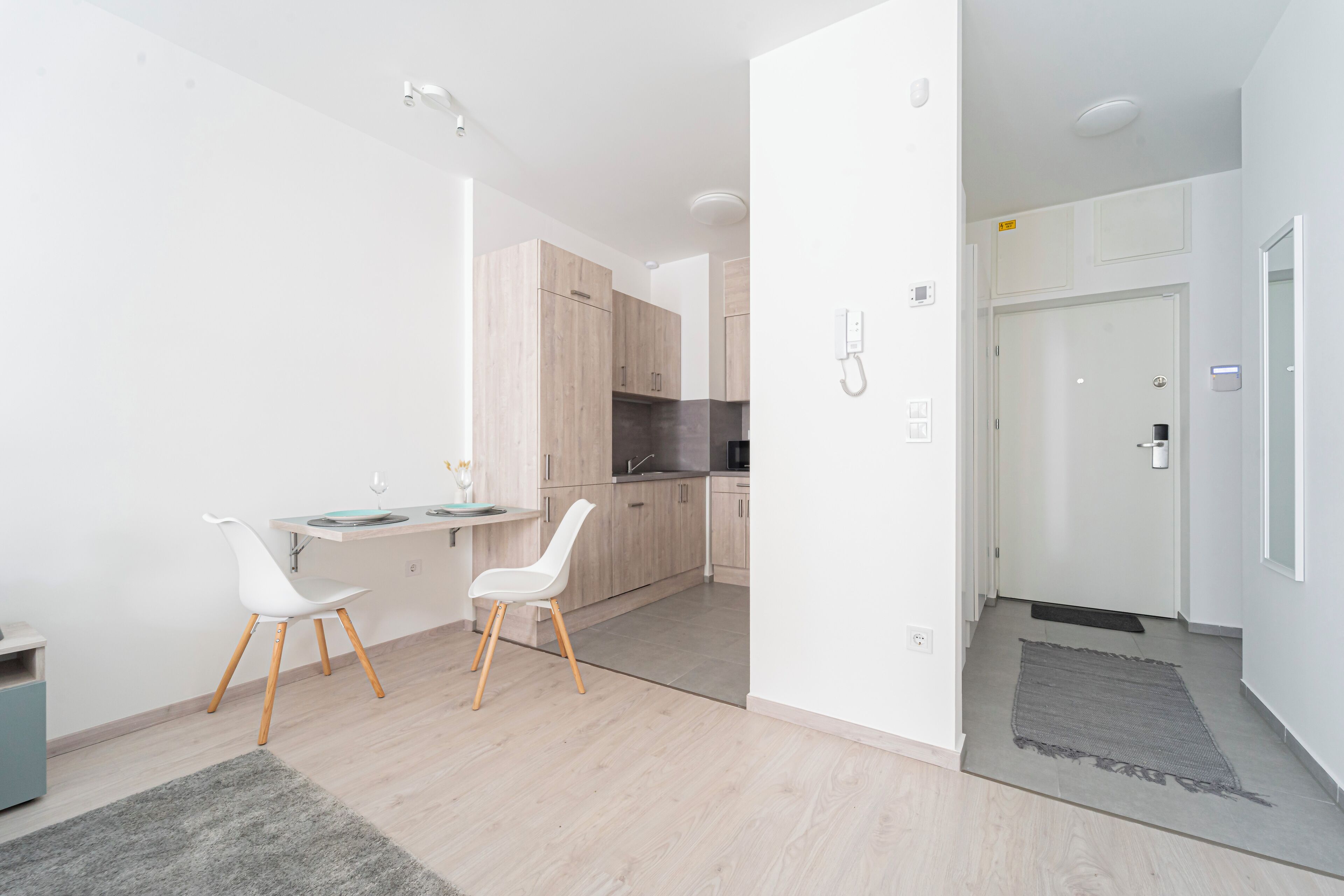A modern kitchen with wooden cabinets, a dining area with white chairs, and a grey rug on the floor.