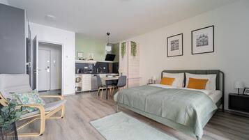 A modern bedroom with a bed, a desk, and a chair. There is a kitchenette in the background.