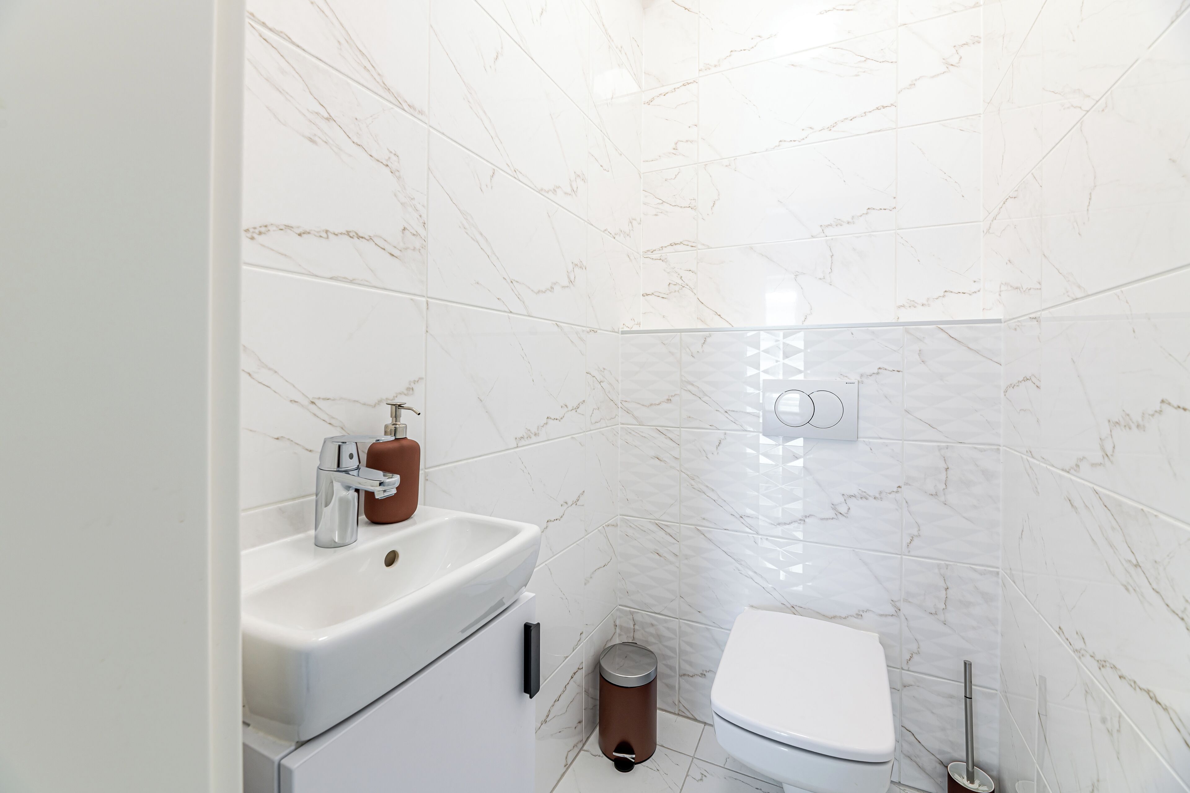 A modern bathroom with white marble tiles, a white sink with a soap dispenser, a toilet, and a trash bin.