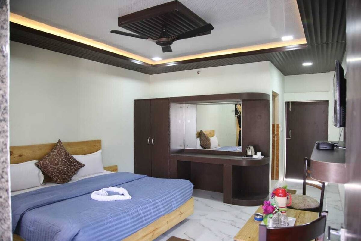 Habitación de hotel con cama, escritorio, espejo, televisor y ventilador de techo.