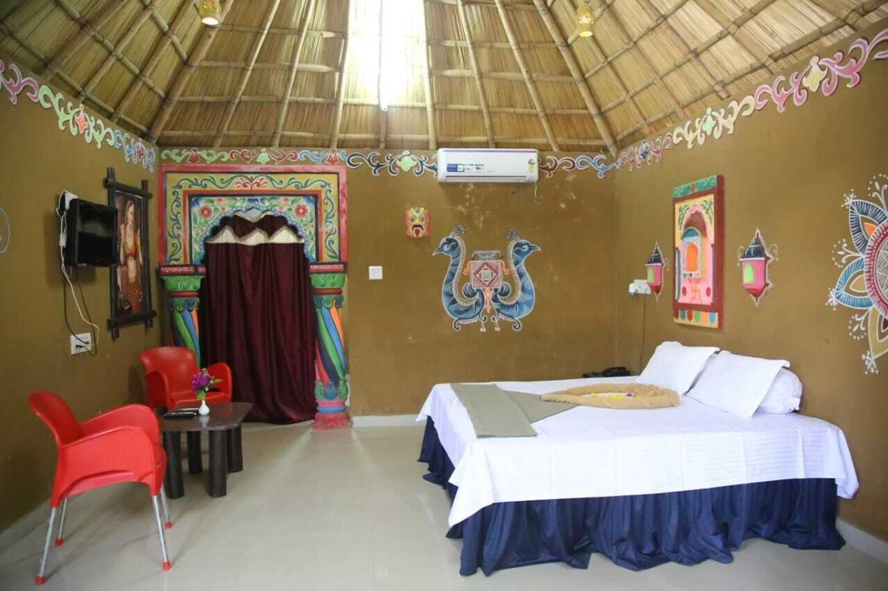 A room with a bed, a table, and a chair. There is a decorative wall with colorful patterns and a thatched roof.