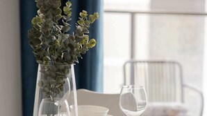 Mesa de jantar posta com pratos brancos, tigelas, talheres e um vaso de vidro transparente com folhagem verde.