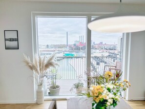 A modern living room with a large window overlooking a cityscape, including a river and industrial buildings. There are potted plants on the windowsill and a framed picture on the wall. - Modern Apartment with a View

 (København)