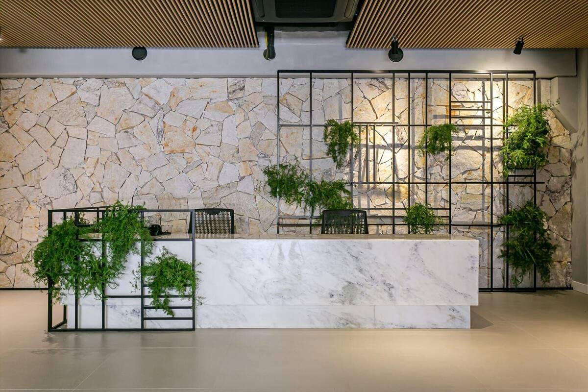 Un comptoir d’accueil avec un mur en pierre en arrière-plan, des étagères en métal ornées de plantes et un comptoir en marbre.