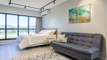 A modern hotel room with a bed, a gray sofa, a small side table, and large windows offering a view of the outdoors.