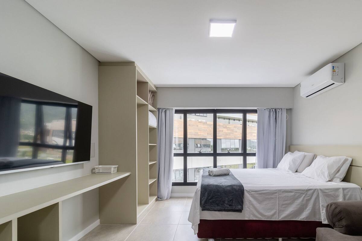 Une chambre d’hôtel avec un lit, un téléviseur posé sur une étagère, une fenêtre avec des rideaux et un appareil de climatisation.