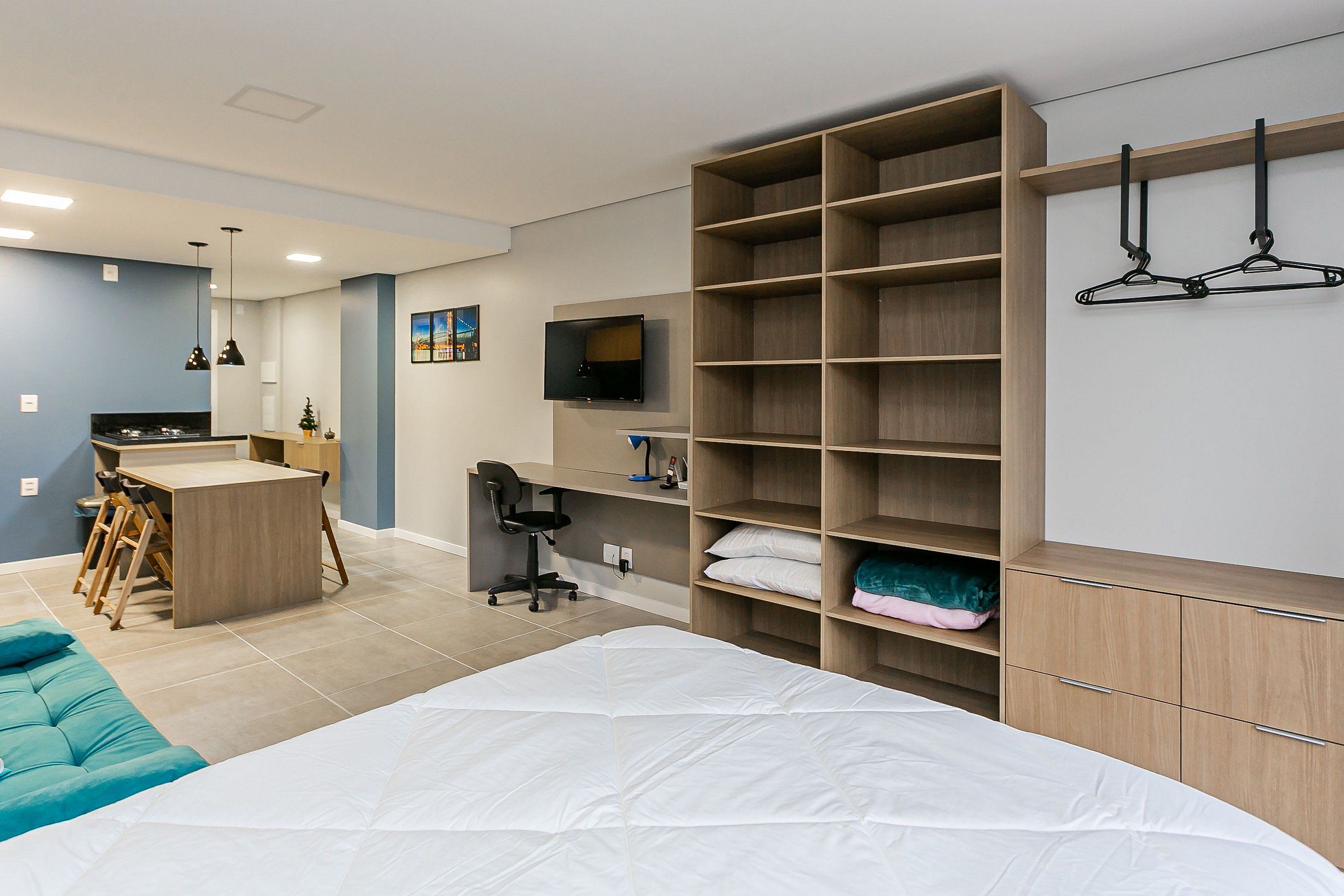 A modern bedroom with a bed, a desk, and a chair. There is a TV mounted on the wall and a wardrobe with shelves.