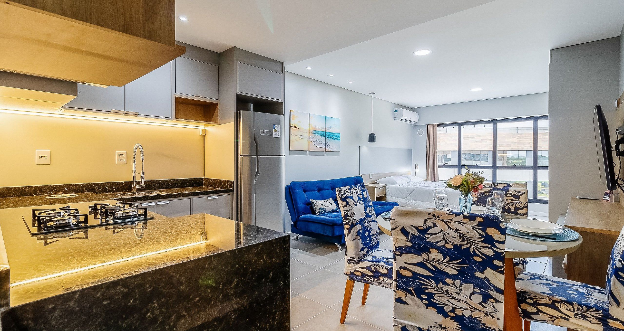 A modern kitchen with a built-in stove, a refrigerator, and a dining area with a blue sofa and floral chairs.