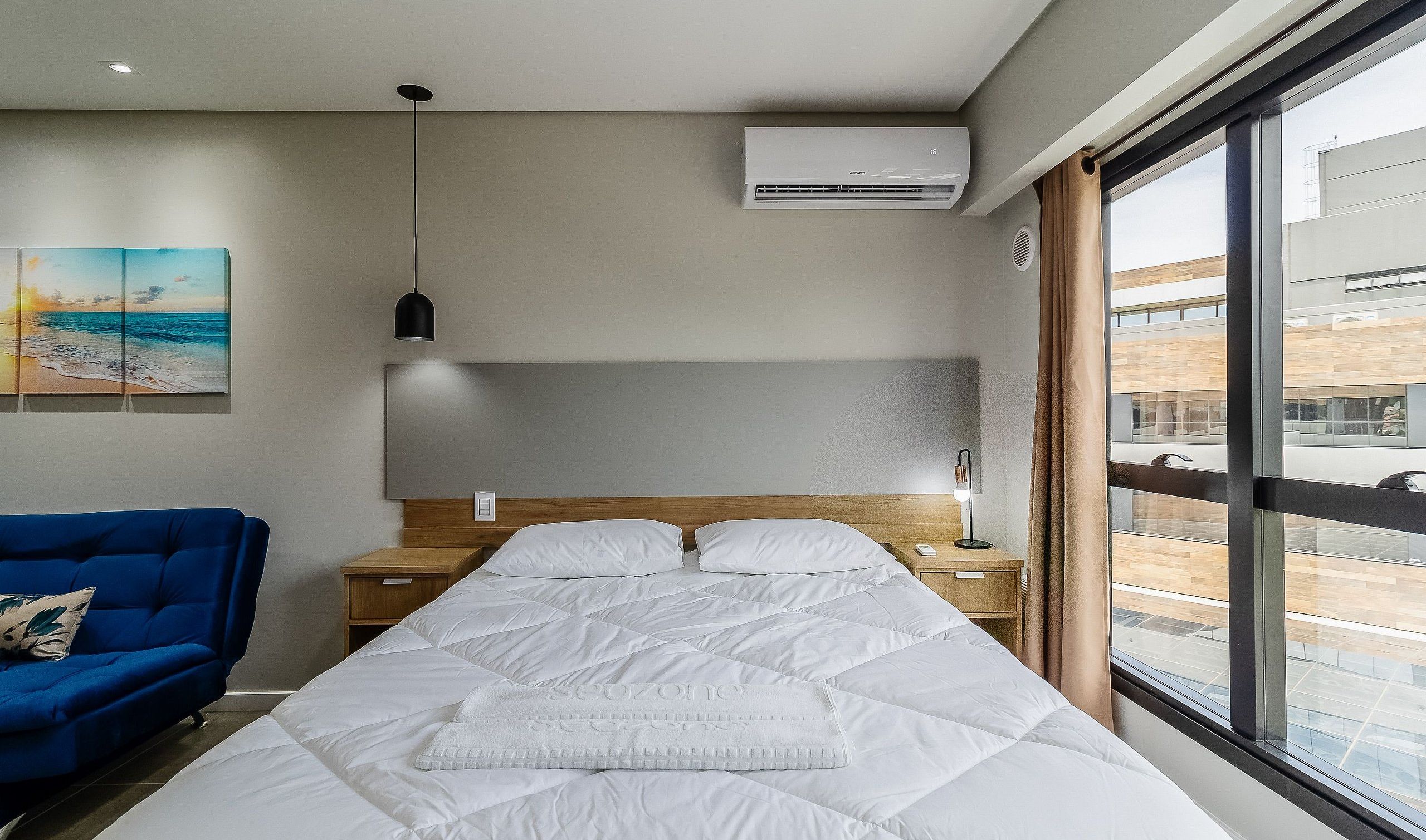 Una habitación de hotel con cama, mesita de noche, lámpara, una silla azul y un ventanal grande.