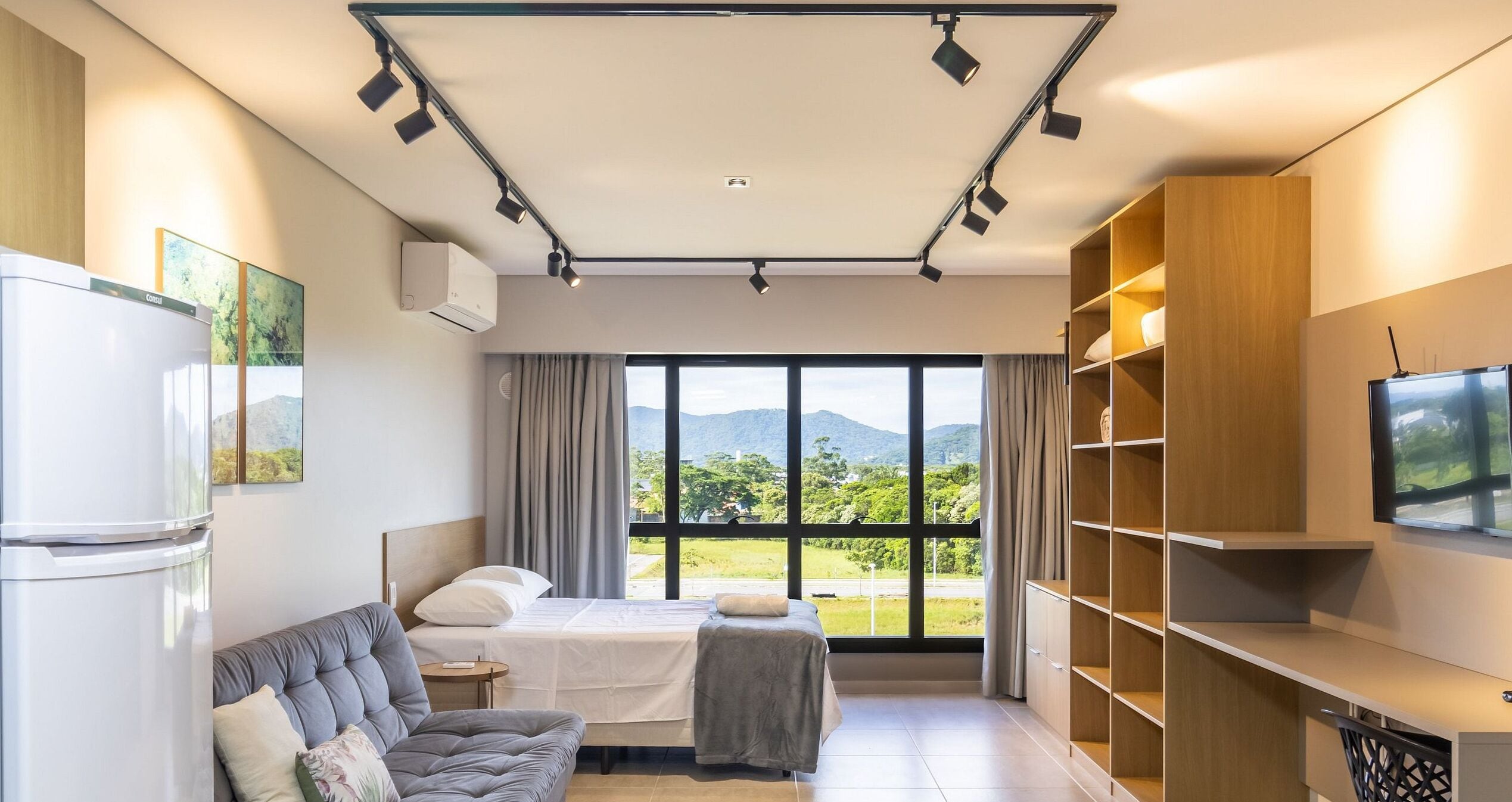 A modern hotel room with a bed, a sofa, a TV, and a large window offering a view of the outdoors.