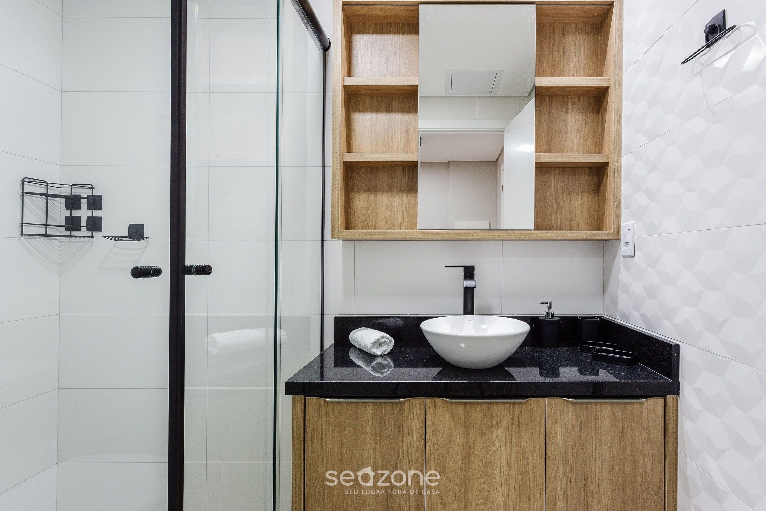 Un baño moderno con cabina de ducha de vidrio, tocador de madera con encimera negra, lavabo blanco y espejo sobre el tocador.