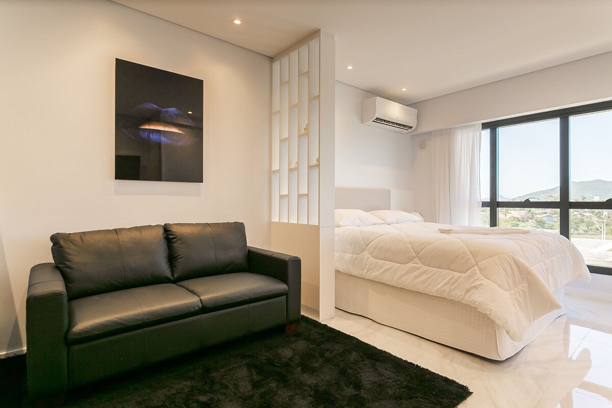 A modern bedroom with a black leather sofa, a white bed, a large window, and an air conditioning unit.