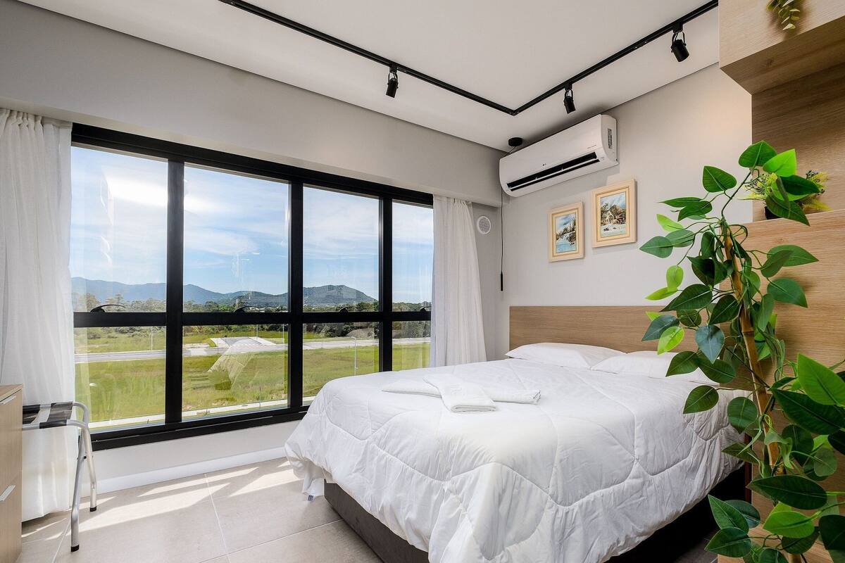 Une chambre avec une grande fenêtre, un lit, un climatiseur et une plante.