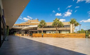 Een modern gebouw met een plat dak, grote ramen en een geplaveid plein met bankjes.