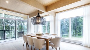 Ett rymligt matområde med ett träbord, sex stolar och en hängande lampa. Stora fönster ger utsikt över ett skogsområde.
