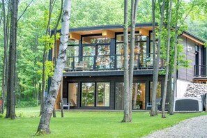 A modern two-story house with large windows and a balcony, surrounded by trees.