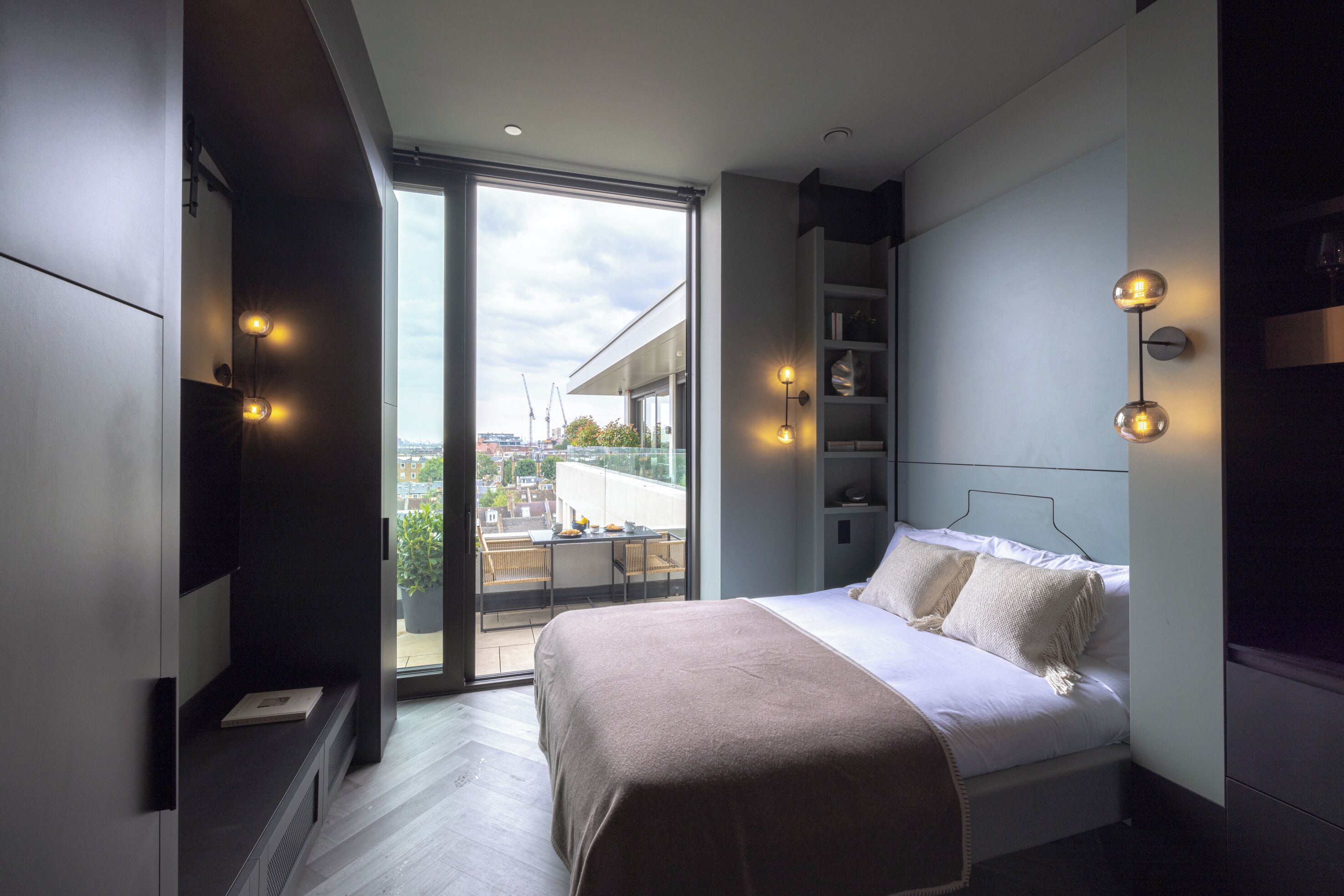 A modern bedroom with a large bed, built-in shelving, and a view of the city through a large window.