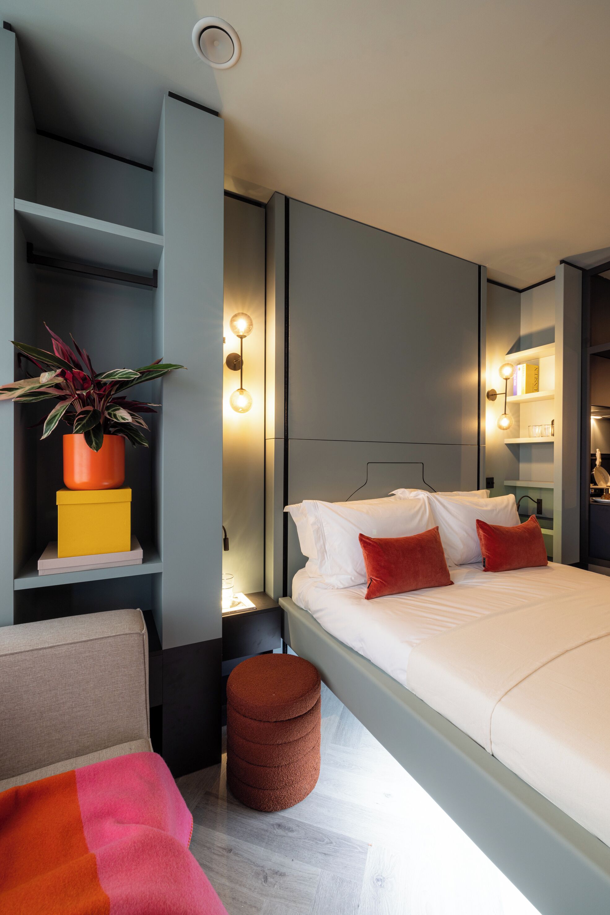 A modern hotel room with a bed, bedside tables, a small stool, a chair, a shelf with a plant, and a mirror.