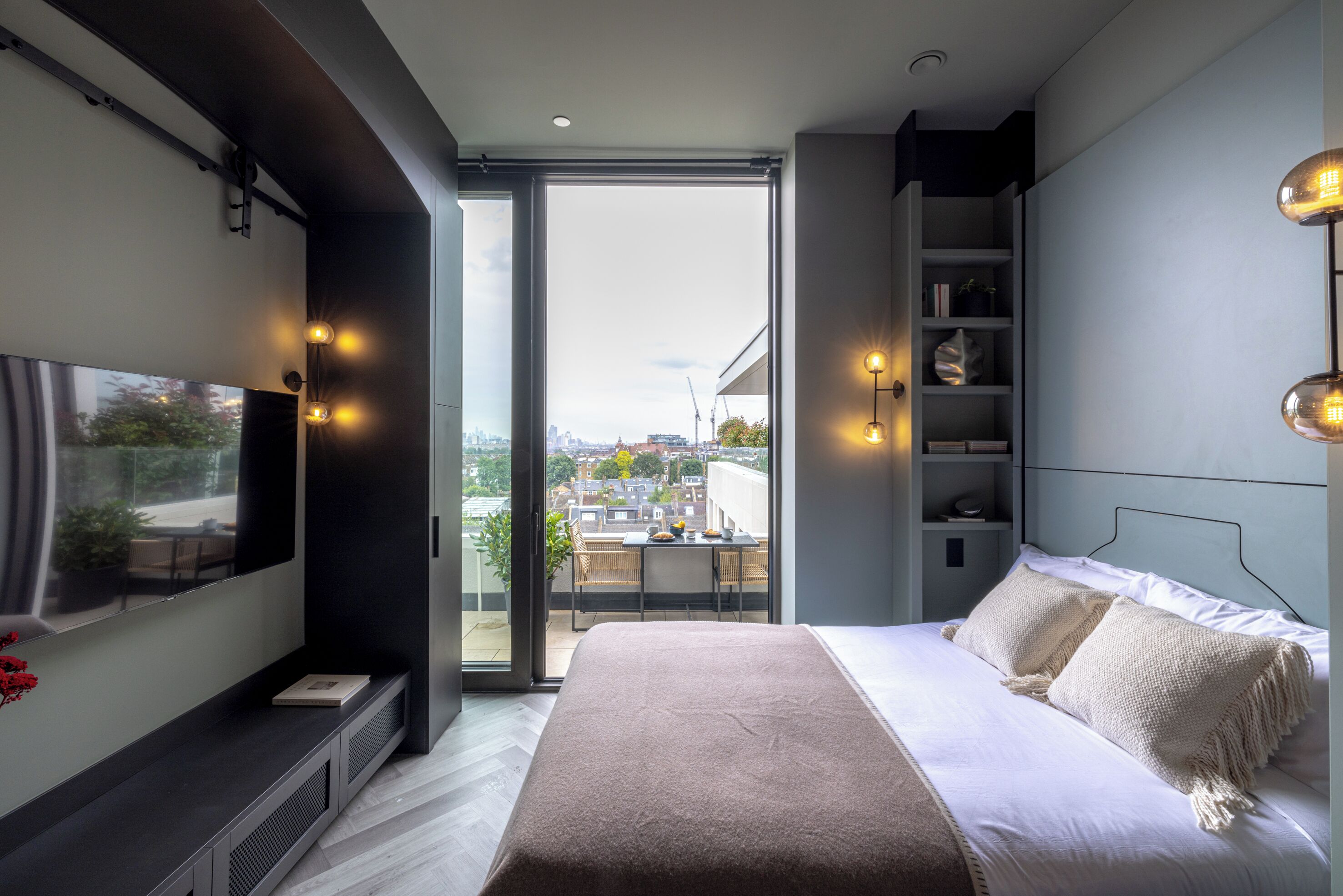 A modern bedroom with a large bed, a TV on the wall, and a balcony with a city view.