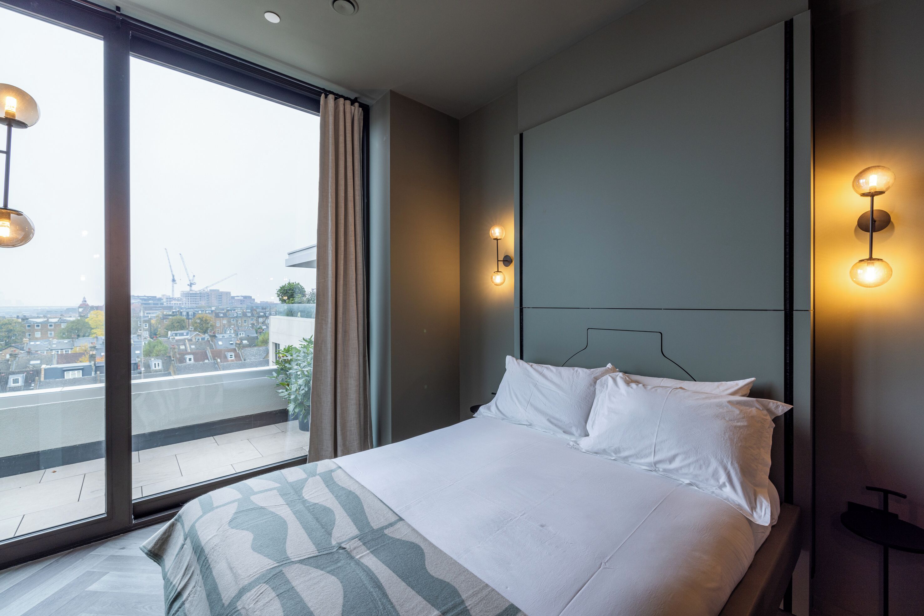 A modern hotel room with a large window, a bed with white linens, and wall-mounted lighting.