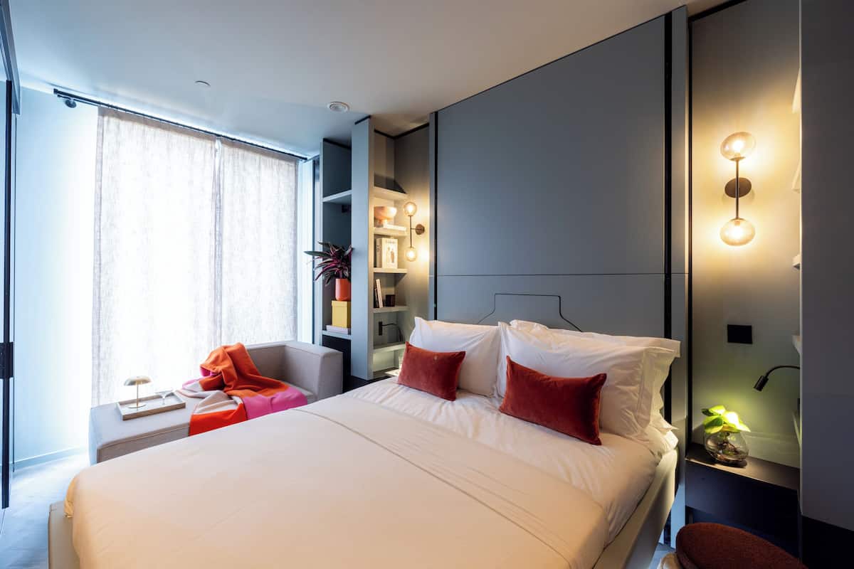 A modern hotel room with a large bed, a bedside table with a lamp, a chair, and a built-in shelf with decorative items.