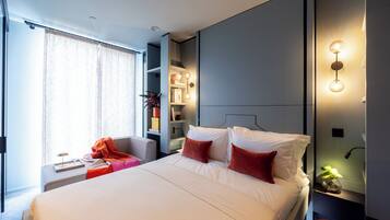 Une chambre d’hôtel moderne dotée d’un grand lit, d’une table de chevet avec une lampe, d’une chaise et d’une étagère intégrée où sont disposés des objets décoratifs.