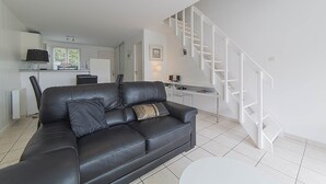 A modern living room with a black leather sofa, a white staircase, and a kitchen in the background. - Maison Verte 4 Pers (LA FLOTTE)