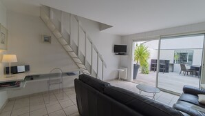 A modern living room with a black leather sofa, a small round table, a TV mounted on the wall, a staircase, and a large sliding glass door leading to an outdoor patio area with a table and chairs. - Maison Verte 4 Pers (LA FLOTTE)