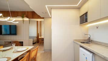 Cozinha moderna com micro-ondas embutido, pia e máquina de lavar louça. Há uma área de jantar com mesa e cadeiras, e uma televisão fixada na parede.