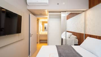 Una habitación de hotel moderna con televisión de pantalla plana, cama con sábanas blancas, manta gris, cabecera de madera y vestidor con espejo.