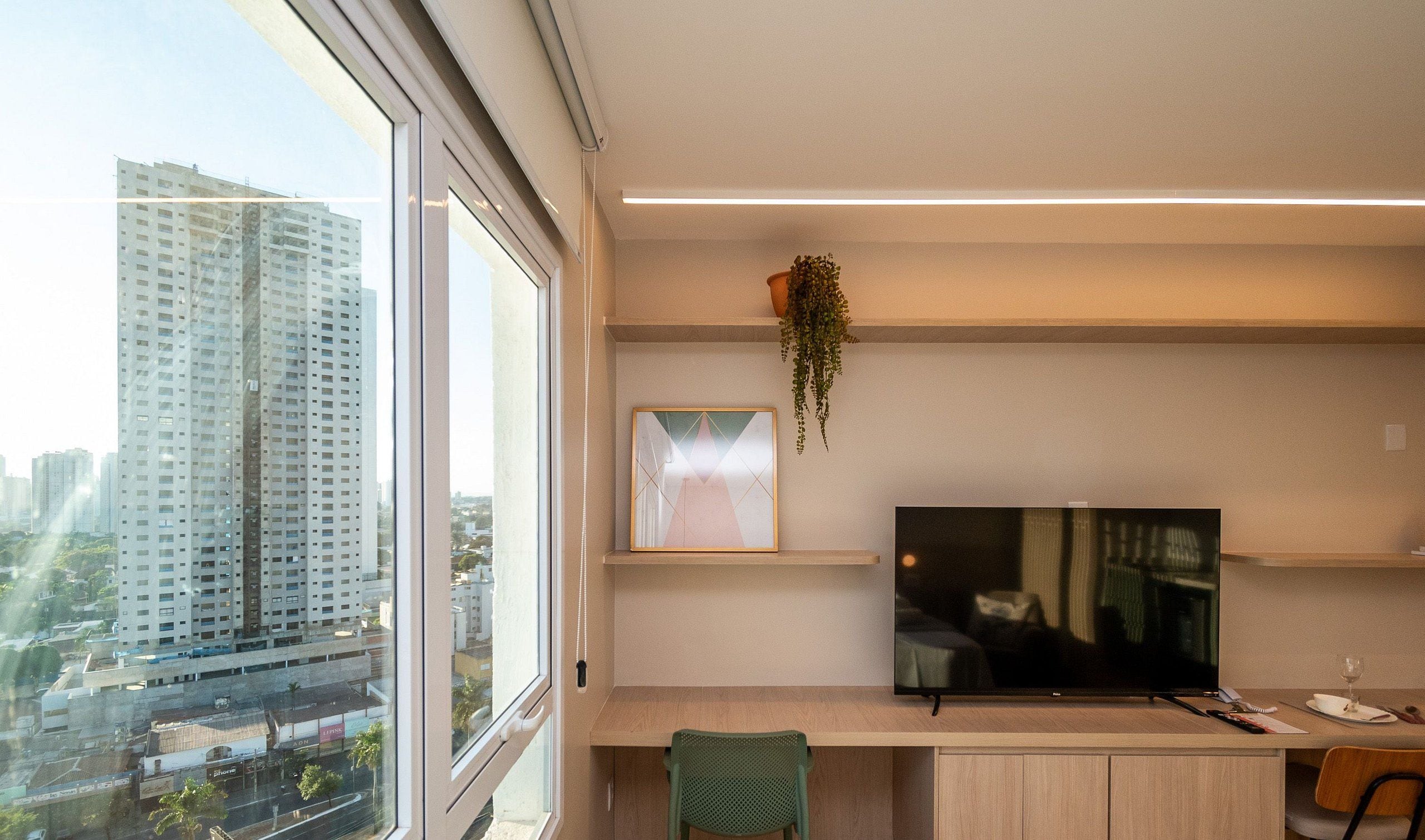 A modern office space with a large window, a desk, a chair, a television, and a hanging plant.