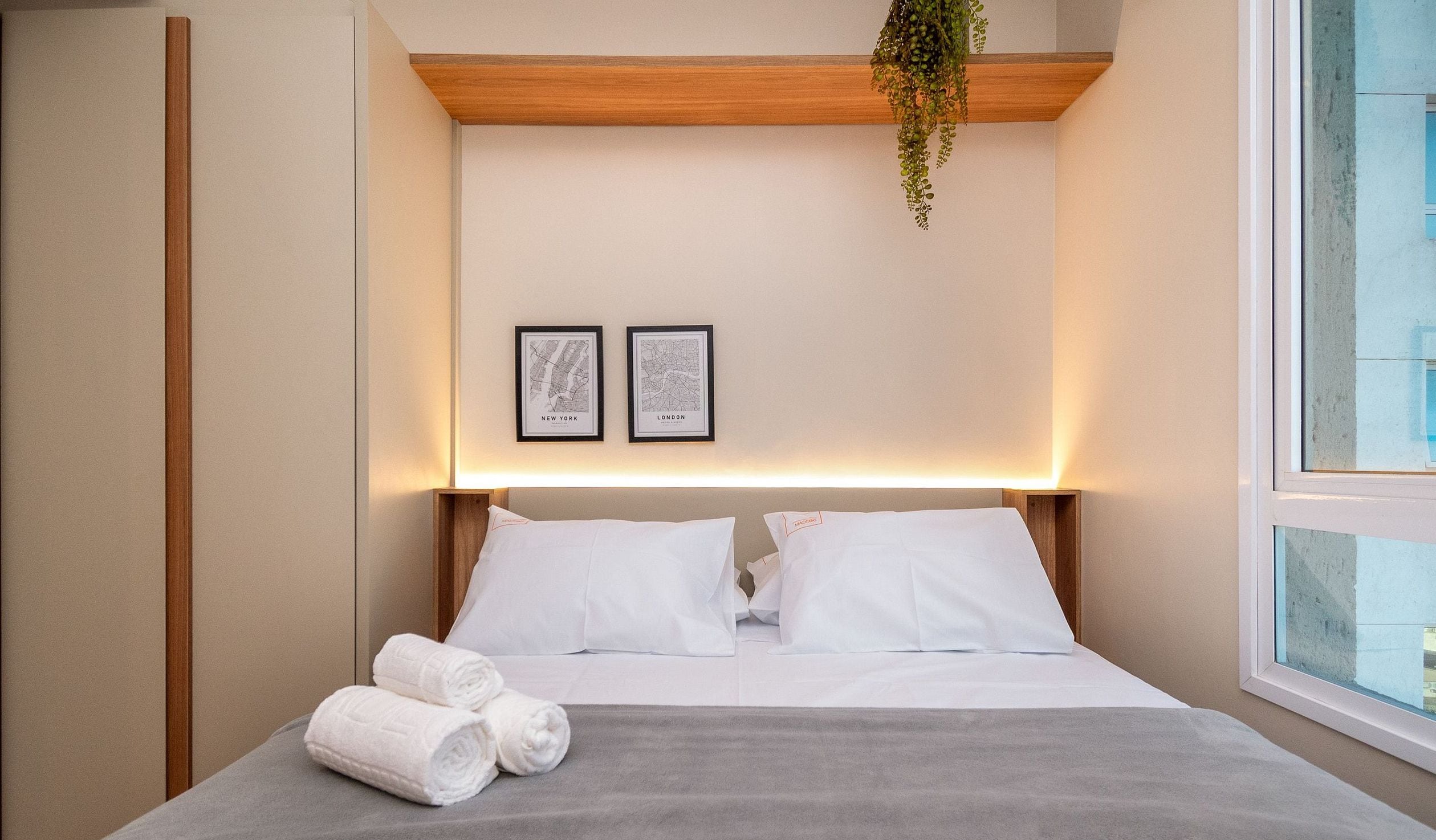 Una habitación de hotel con una cama, dos mapas enmarcados, una planta colgante y una ventana.