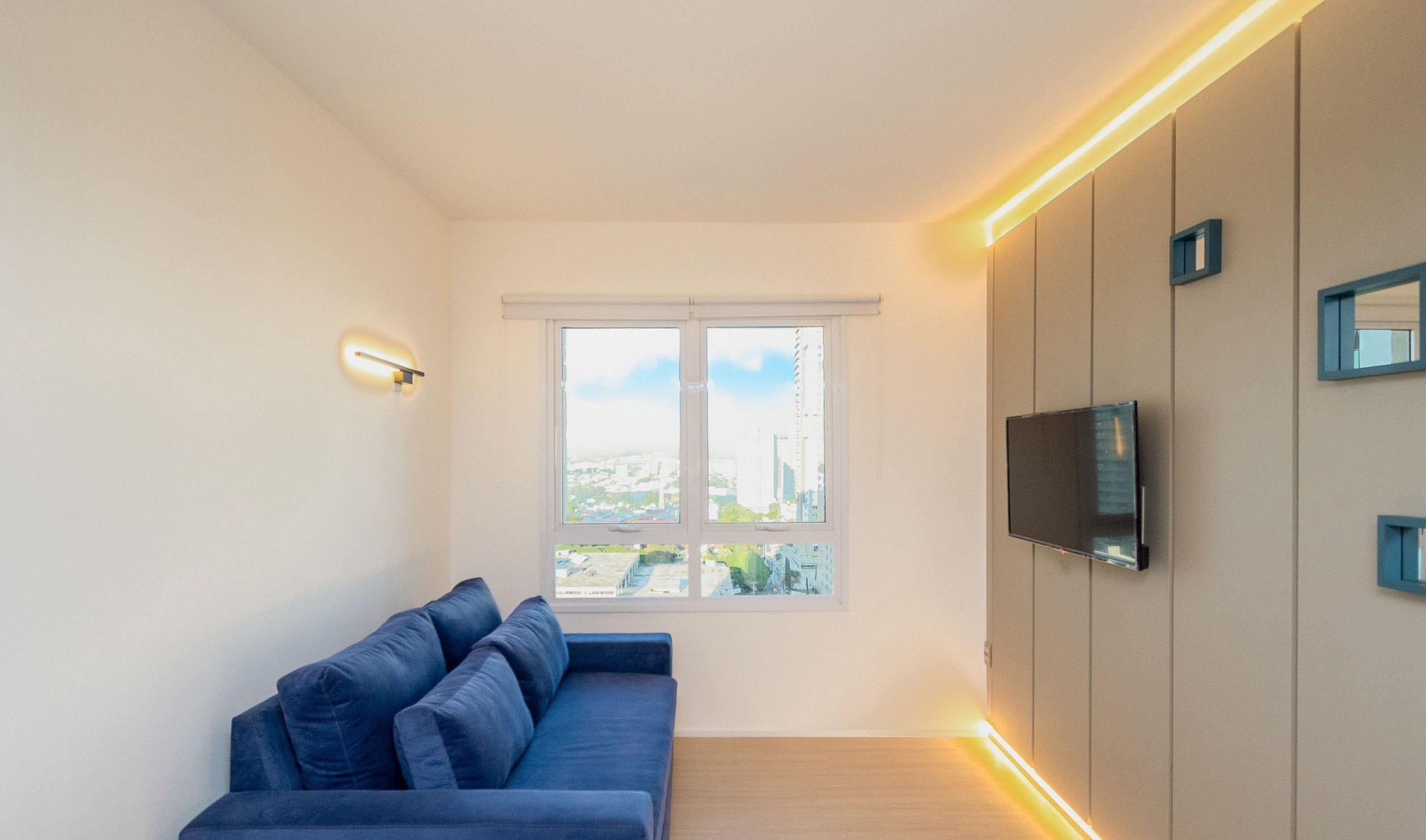 A modern living room with a sofa, a TV mounted on the wall, and a window with a city view.