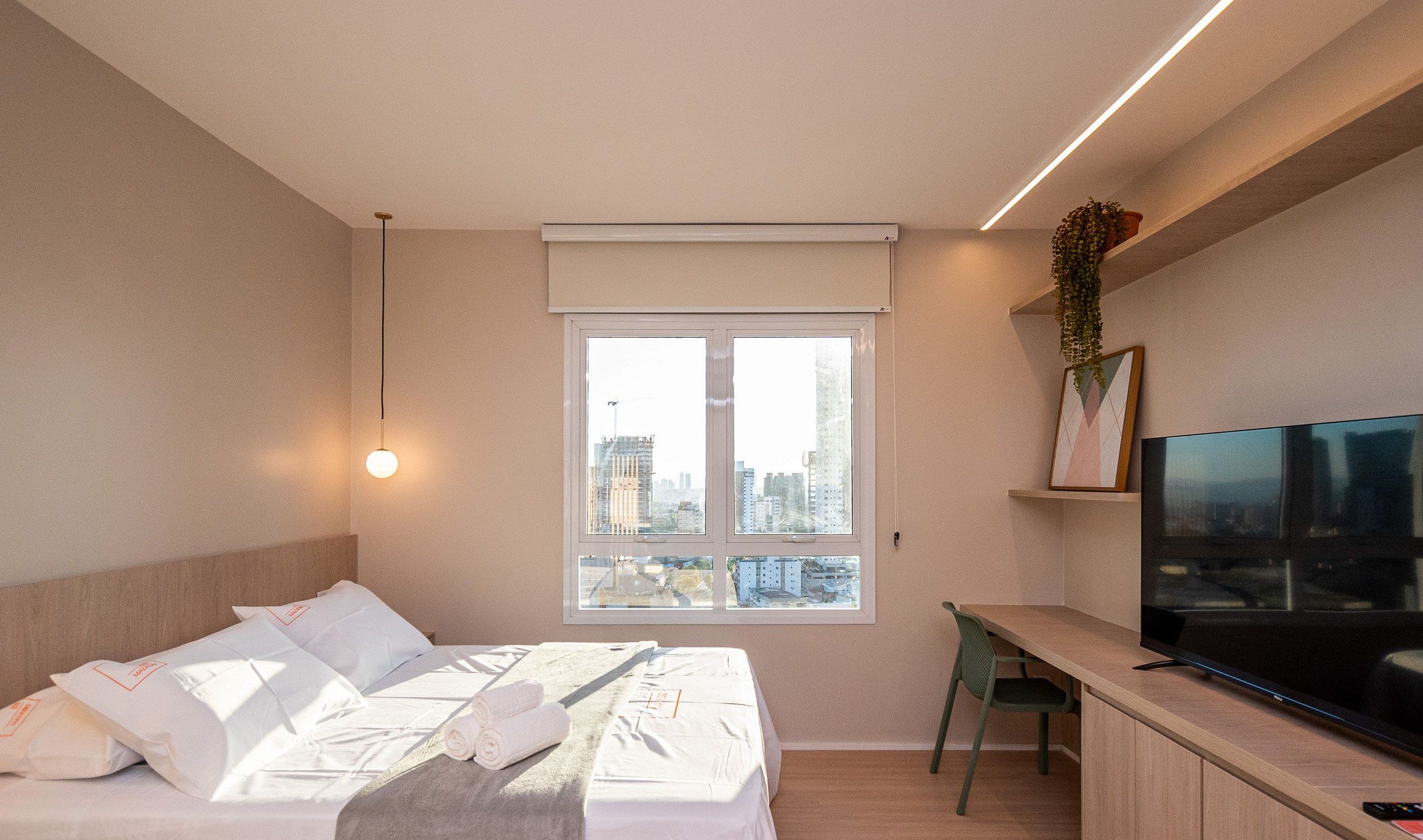 A modern bedroom with a bed, a desk, a television, and a window with a city view.