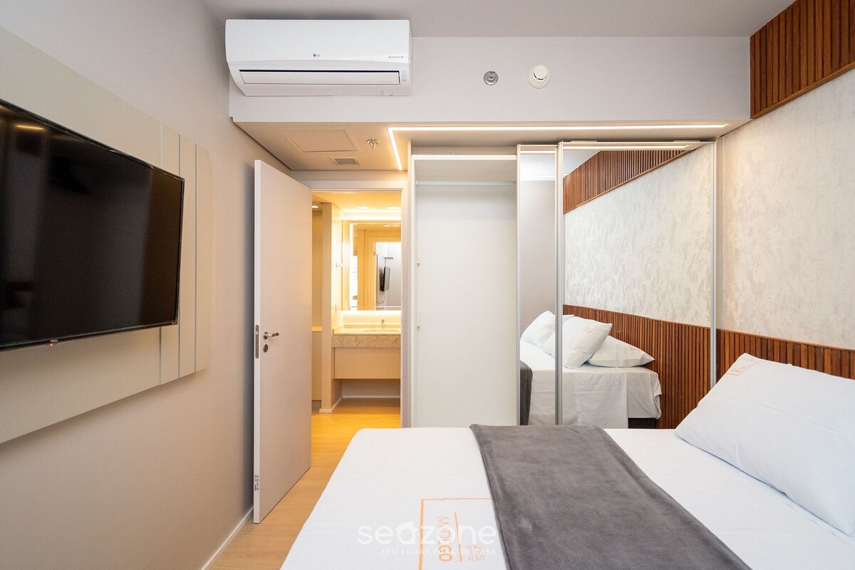 Une chambre d’hôtel moderne dotée d’une télévision à écran plat, d’un lit avec du linge de maison blanc, d’un plaid gris, d’une tête de lit en bois et d’un dressing avec un miroir.