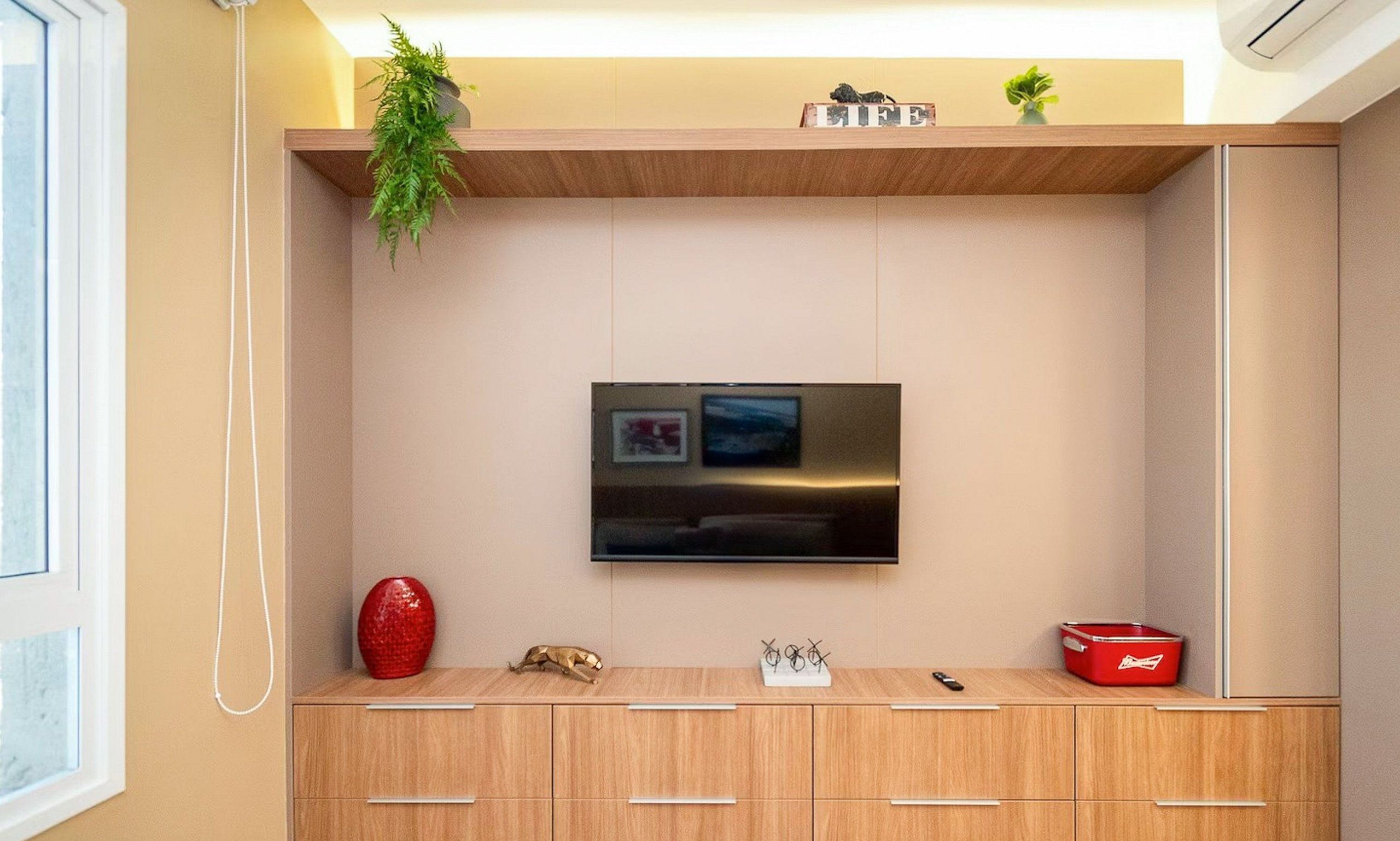 Uma sala de estar moderna com televisão de tela plana na parede, armários de madeira abaixo e um pequeno vaso vermelho à esquerda.