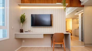 Une chambre d’hôtel moderne avec un téléviseur à écran plat fixé à un mur blanc, un plafond en bois, un petit bureau avec une chaise et une plante en pot.