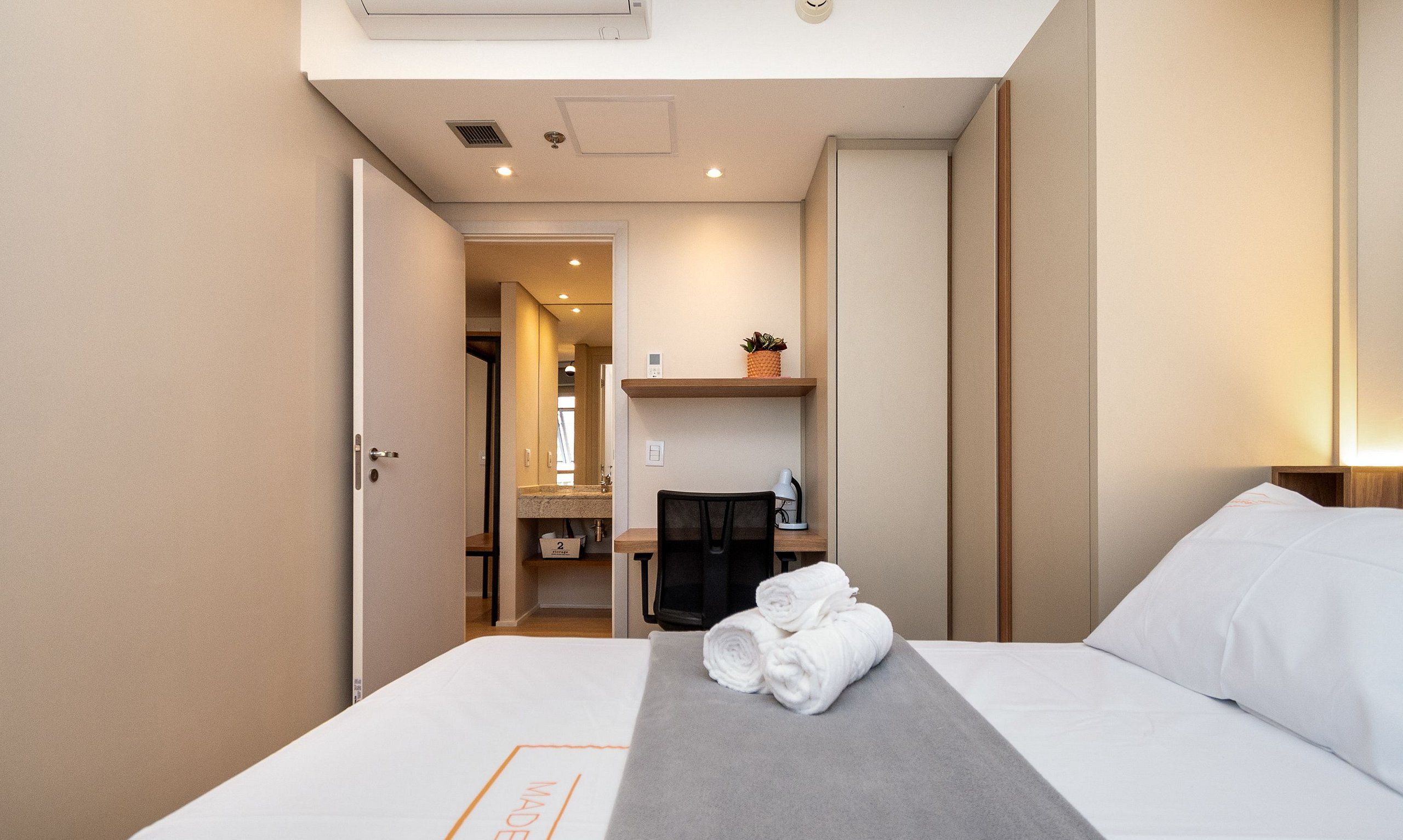 Una camera d'albergo con un letto, una scrivania, una sedia, una piccola pianta e un bagno visibile attraverso una porta aperta.