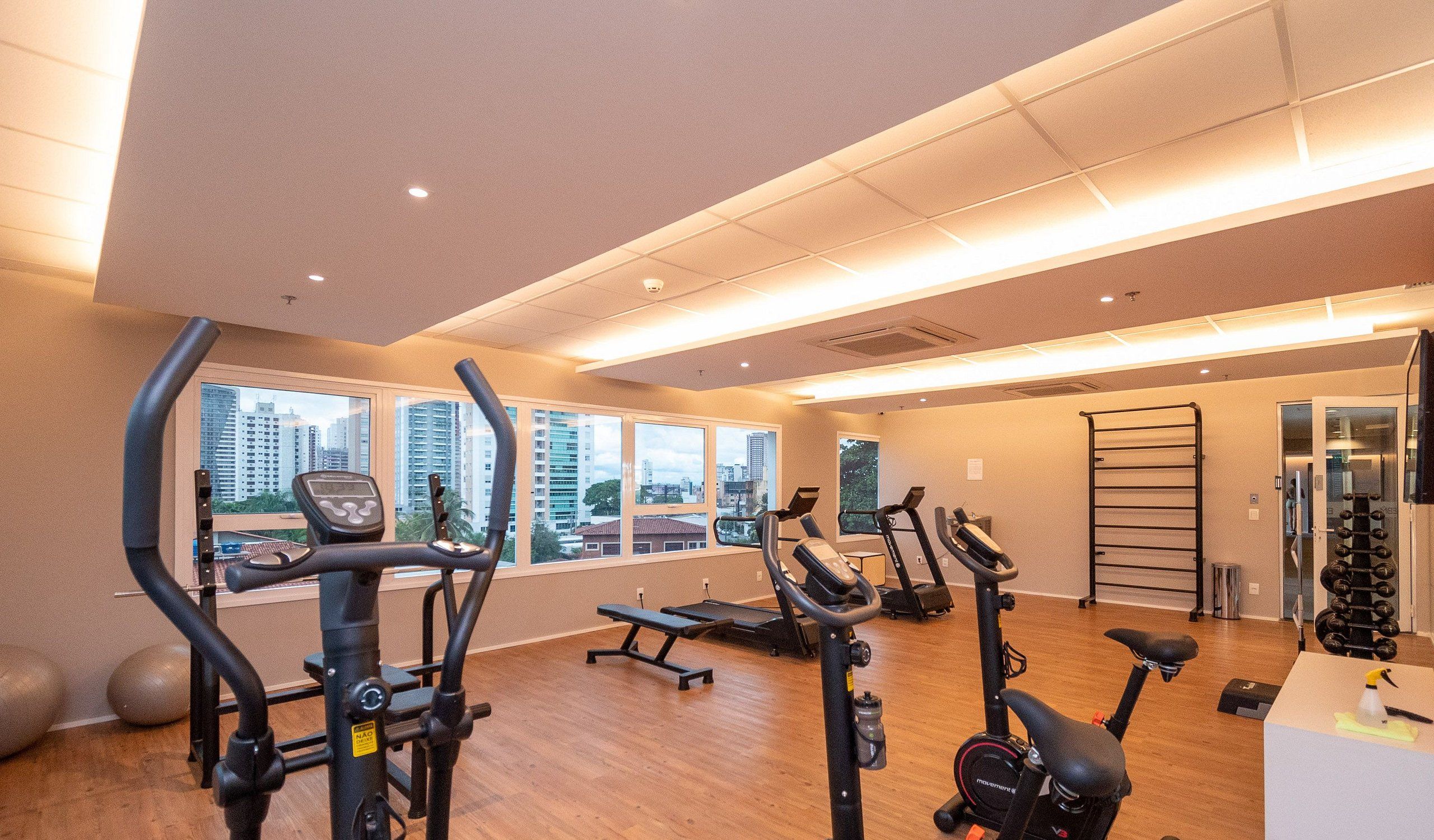 Un gimnasio con equipo de ejercicio diverso, amplias ventanas y vista a edificios.